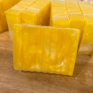 May include: Close-up of several rectangular bars of handmade soap. The soap is a vibrant yellow color with a pearlescent, swirled design. The bars are stacked together, suggesting a collection or display of the product.