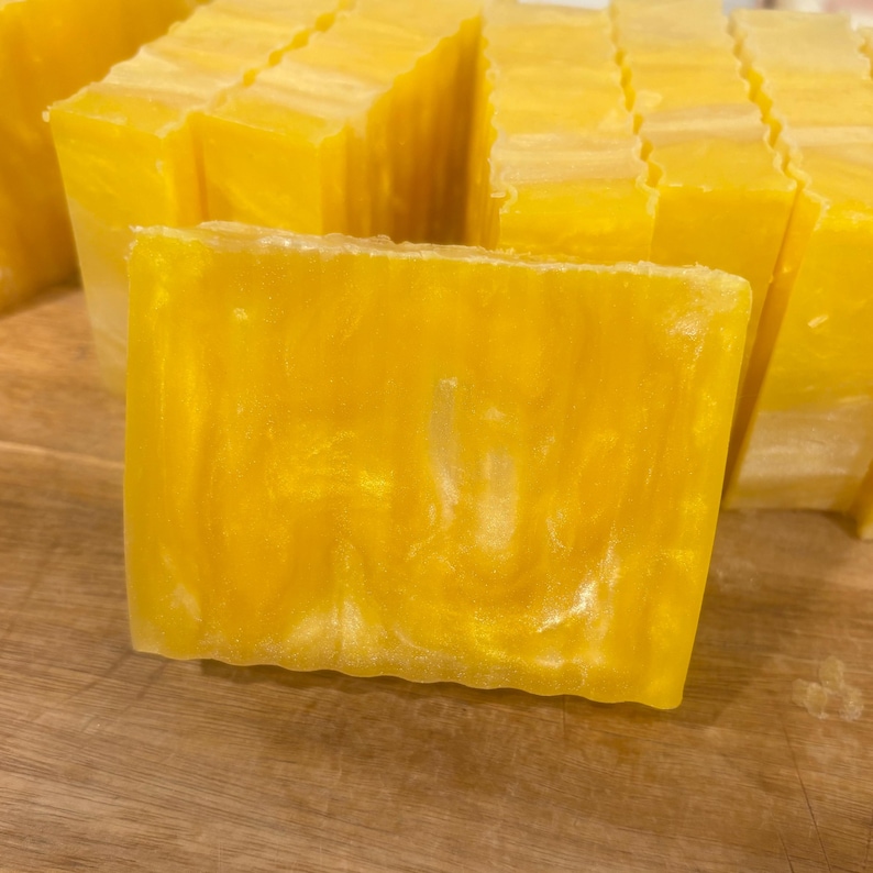 May include: Close-up of several rectangular bars of handmade soap. The soap is a vibrant yellow color with a pearlescent, swirled design. The bars are stacked together, suggesting a collection or display of the product.