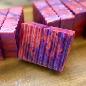Pu&ograve; includere: Una saponetta rettangolare caratterizzata da un motivo marmorizzato con vortici di rosa vivace e viola intenso. La superficie del sapone presenta una finitura testurizzata e scanalata con linee verticali sul davanti e un aspetto lucido.