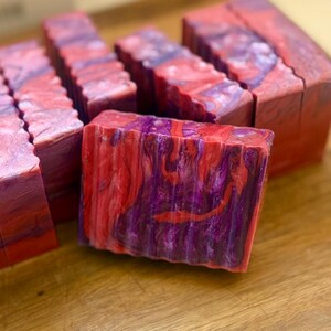 Pu&ograve; includere: Diverse saponette rettangolari fatte a mano caratterizzate da un motivo a vortice marmorizzato in tonalit&agrave; vivaci di viola intenso, rosso brillante e rosa tenue. Ogni saponetta presenta un bordo superiore ondulato e una forma solida.
