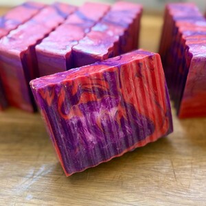 Pu&ograve; includere: Una saponetta rettangolare caratterizzata da un motivo a vortice marmorizzato nei colori viola vibrante e rosa acceso. La superficie del sapone ha una consistenza liscia con striature visibili sul lato, e diverse saponette identiche sono disposte dietro.