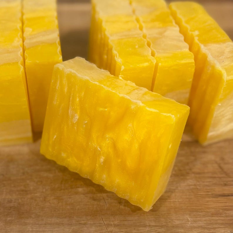 May include: Close-up of several rectangular bars of yellow soap. The soap has a textured surface and a slightly translucent appearance. The bars are arranged on a wooden surface, suggesting a natural or handmade product.