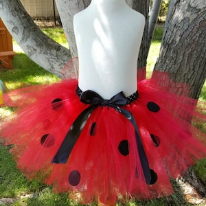May include: Red tulle tutu skirt with black polka dots and a black satin bow. The skirt is on a white mannequin and is displayed against a green grassy background.