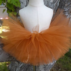 May include: A brown tulle tutu skirt on a white mannequin. The skirt is made of multiple layers of tulle and has a gathered waistband.