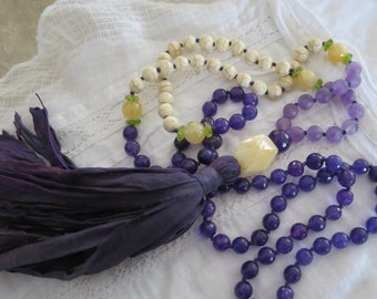 Op de afbeelding: Een paarse en witte kralenketting met een kwast en een gele stenen hanger. De ketting is gemaakt van amethist, witte howliet en groene aventurijn kralen.