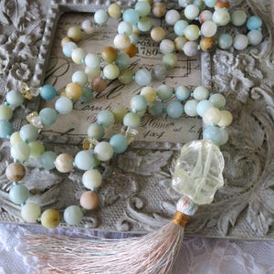 Puede incluir: Un collar largo hecho de cuentas de color azul claro, verde y amarillo con un dije de cristal amarillo claro y una borla rosa claro y azul. El collar se muestra en un marco gris adornado con una inscripci&oacute;n vintage.