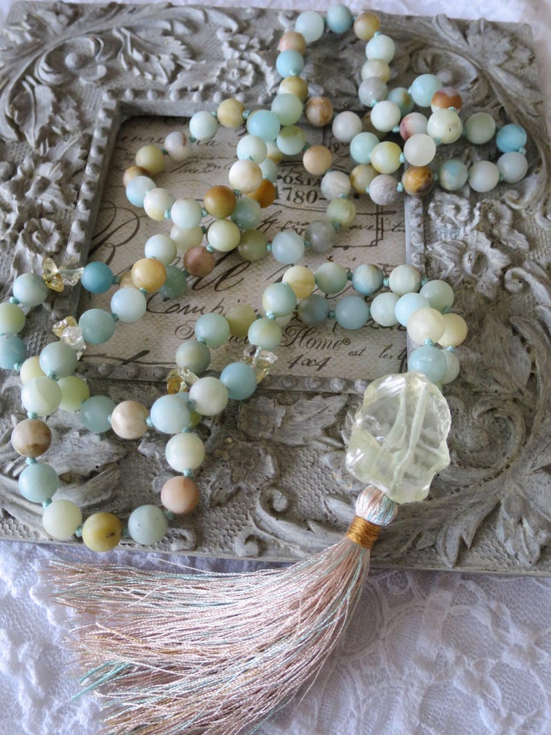 Puede incluir: Un collar largo hecho de cuentas de color azul claro, verde y amarillo con un dije de cristal amarillo claro y una borla rosa claro y azul. El collar se muestra en un marco gris adornado con una inscripci&oacute;n vintage.