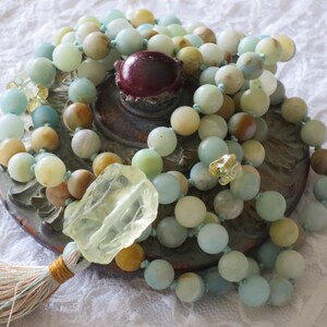 Puede incluir: Un collar mala de m&uacute;ltiples hilos hecho con cuentas de color azul claro, verde y marr&oacute;n. El collar tiene un borla y un gran dije de cristal transparente.