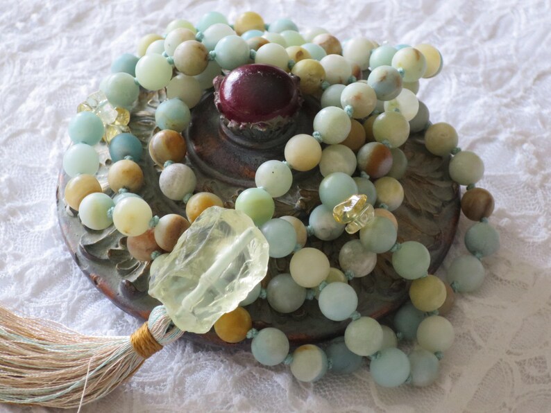 Puede incluir: Un collar mala de m&uacute;ltiples hilos hecho con cuentas de color azul claro, verde y marr&oacute;n. El collar tiene un borla y un gran dije de cristal transparente.