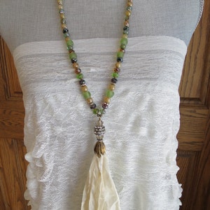 Op de afbeelding: Een lange kralenketting met een cr&egrave;mekleurige kwast. De kralen zijn groen, bruin en goud. De ketting ligt op een witte kant achtergrond.