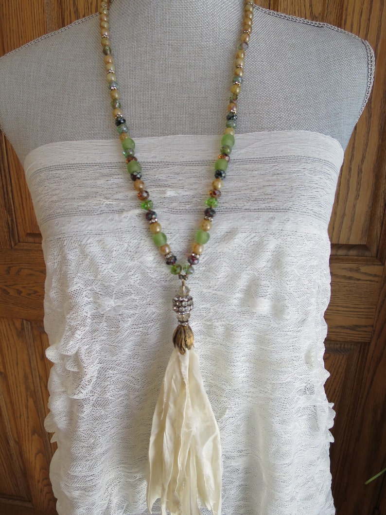 Op de afbeelding: Een lange kralenketting met een cr&egrave;mekleurige kwast. De kralen zijn groen, bruin en goud. De ketting ligt op een witte kant achtergrond.