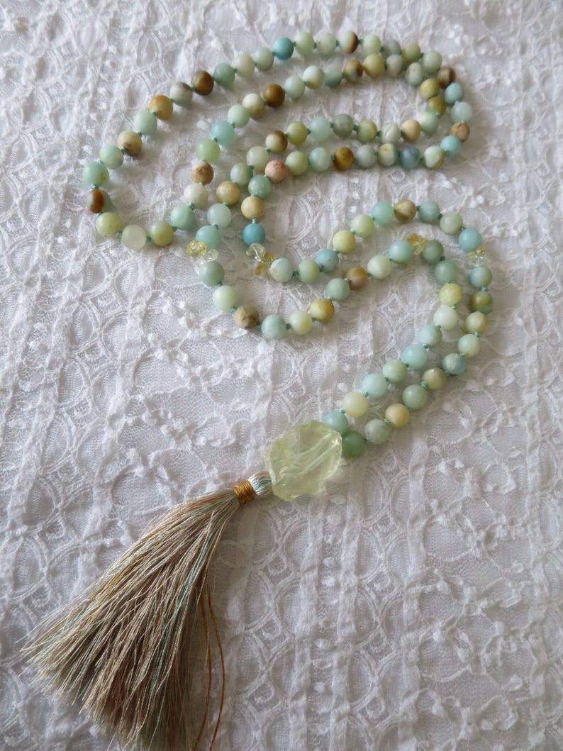 Puede incluir: Un collar mala verde y marr&oacute;n con un borla. El collar est&aacute; hecho de cuentas y tiene una piedra verde grande al final.