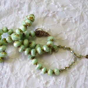 Op de afbeelding: Een vintage ketting met lichtgroene, facetgeslepen kralen en bruine accenten. De ketting heeft een decoratieve sluiting en een goudkleurige bloemhanger. De kralen zijn geregen op een delicate ketting, rustend op een wit kanten oppervlak.