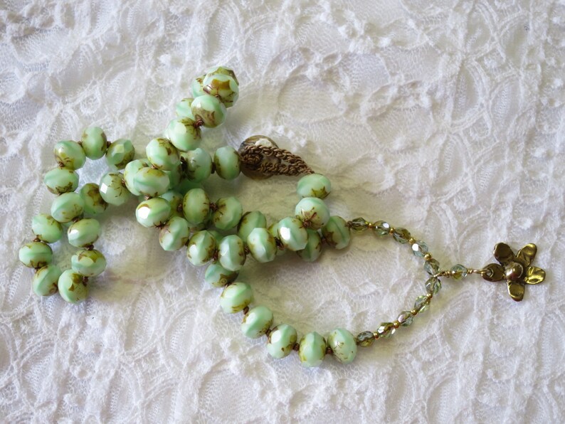 Op de afbeelding: Een vintage ketting met lichtgroene, facetgeslepen kralen en bruine accenten. De ketting heeft een decoratieve sluiting en een goudkleurige bloemhanger. De kralen zijn geregen op een delicate ketting, rustend op een wit kanten oppervlak.