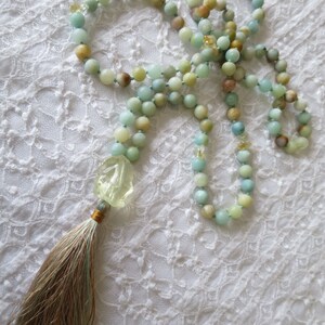 Puede incluir: Un collar mala de piedras preciosas verdes y azules con un colgante de piedra verde tallada y un borla marr&oacute;n. El collar est&aacute; colocado sobre una superficie de encaje blanco.