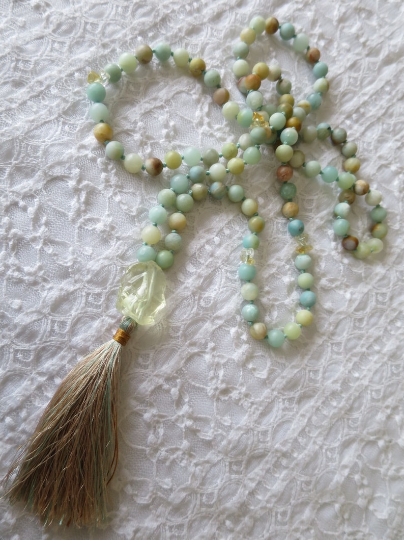 Puede incluir: Un collar mala de piedras preciosas verdes y azules con un colgante de piedra verde tallada y un borla marr&oacute;n. El collar est&aacute; colocado sobre una superficie de encaje blanco.