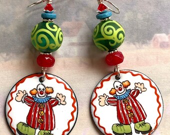 Op de afbeelding: Een paar oorhangers met een groene en witte swirl-kraal, een rode kraal en een ronde metalen hanger met een cartoon-clownillustratie.