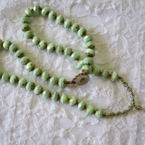 Op de afbeelding: Een ketting met lichtgroene facetkralen met bruine accenten. De ketting is geregen op een bruin koord en heeft een bloemvormige hanger. De kralen zijn in een cirkelvorm gerangschikt.