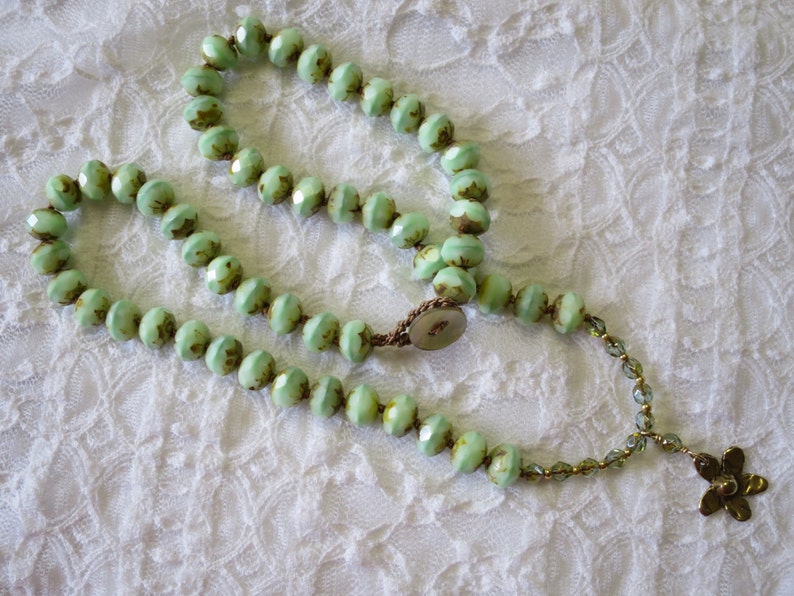 Op de afbeelding: Een ketting met lichtgroene facetkralen met bruine accenten. De ketting is geregen op een bruin koord en heeft een bloemvormige hanger. De kralen zijn in een cirkelvorm gerangschikt.