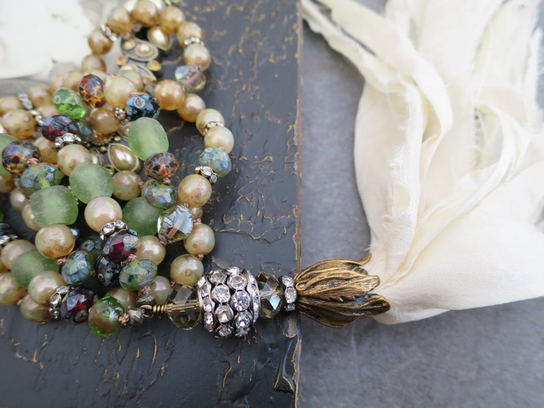Op de afbeelding: Een meerstrengige ketting met groene, bruine en witte kralen, een met kristallen bezette hanger en een gouden kwast.
