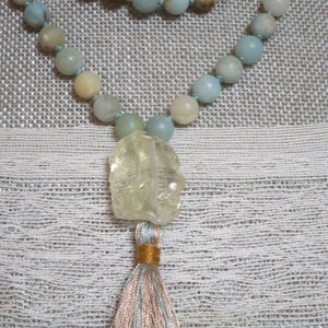 Puede incluir: Un collar mala de color azul claro y beige con un colgante de cuarzo transparente y un borla de hilos azul claro, beige y dorado.