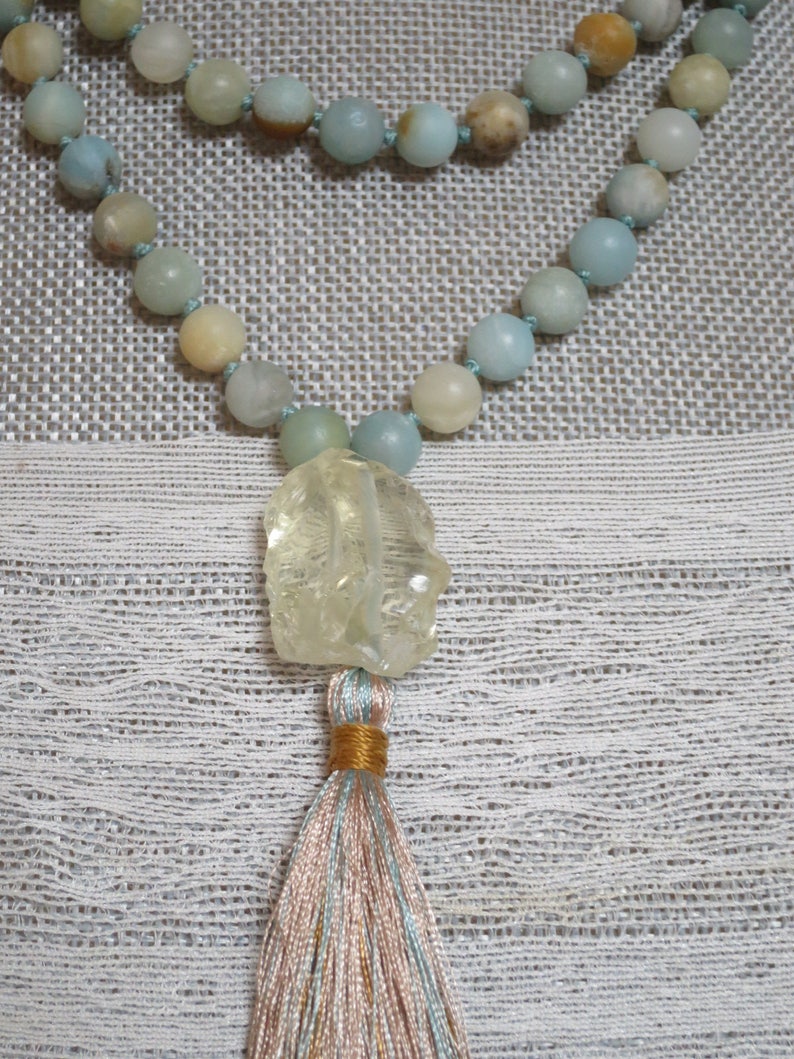 Puede incluir: Un collar mala de color azul claro y beige con un colgante de cuarzo transparente y un borla de hilos azul claro, beige y dorado.