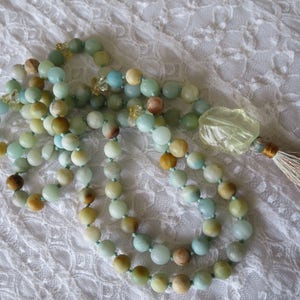 Puede incluir: Un collar de cuentas verdes y marrones con un gran colgante de piedra facetada transparente y un borla. El collar est&aacute; sobre un fondo de encaje blanco.