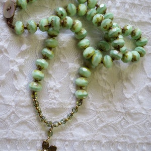 Op de afbeelding: Een vintage-stijl ketting met lichtgroene, facetgeslepen kralen met bruine accenten. De ketting heeft een goudkleurige bloemhanger en een knoopsluiting. De kralen zijn geregen op een bruin koord.