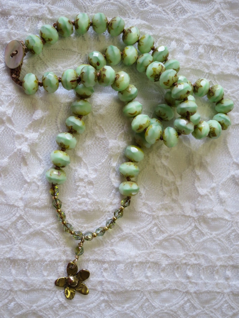 Op de afbeelding: Een vintage-stijl ketting met lichtgroene, facetgeslepen kralen met bruine accenten. De ketting heeft een goudkleurige bloemhanger en een knoopsluiting. De kralen zijn geregen op een bruin koord.