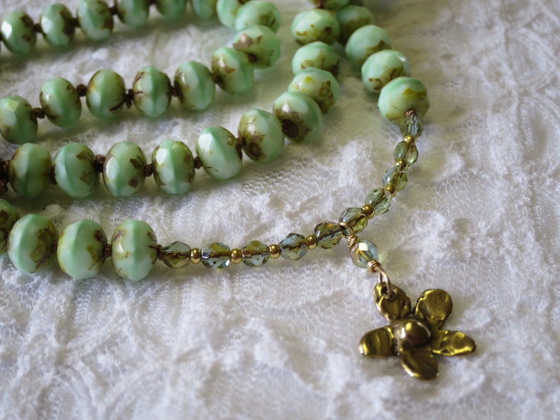 Op de afbeelding: Een close-up van een ketting met lichtgroene, facetgeslepen kralen met bruine accenten. De ketting heeft een goudkleurige bloemhanger en kleinere groene en gouden kralen. De achtergrond is een witte, kanten stof.