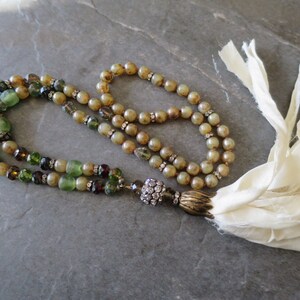 Op de afbeelding: Een lange, meerstrengs ketting met groene, bruine en heldere kralen. De ketting heeft een kwast van witte stof en een hanger met kristallen.