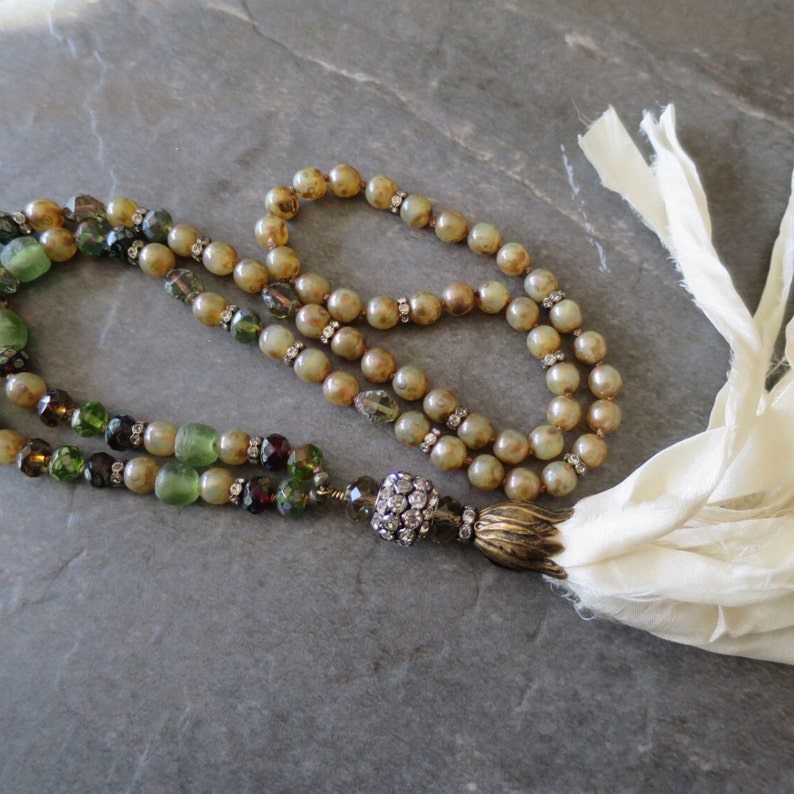 Op de afbeelding: Een lange, meerstrengs ketting met groene, bruine en heldere kralen. De ketting heeft een kwast van witte stof en een hanger met kristallen.