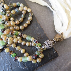 Op de afbeelding: Een meerstrengs ketting met bruine, groene en heldere glazen kralen, gouden accenten en een met kristallen bezette hanger. De ketting wordt getoond op een zwart frame met een witte stoffen kwast.