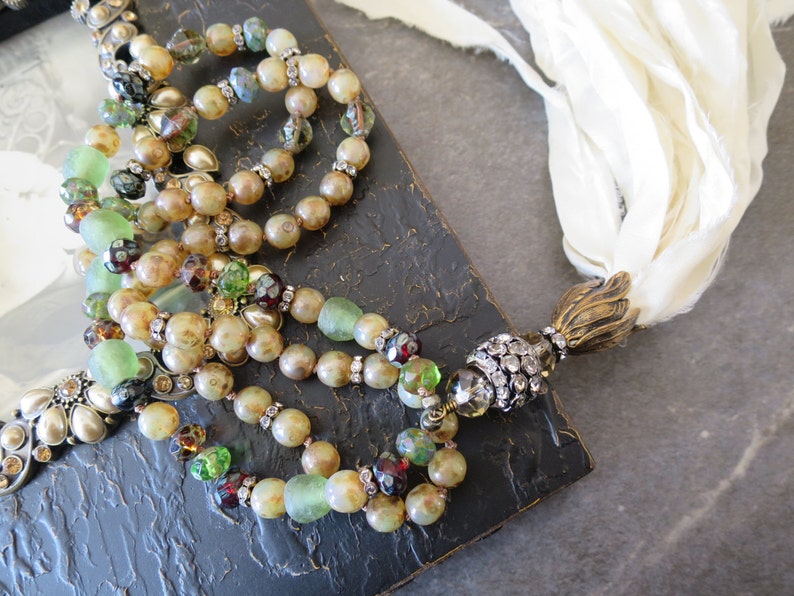 Op de afbeelding: Een meerstrengs ketting met bruine, groene en heldere glazen kralen, gouden accenten en een met kristallen bezette hanger. De ketting wordt getoond op een zwart frame met een witte stoffen kwast.