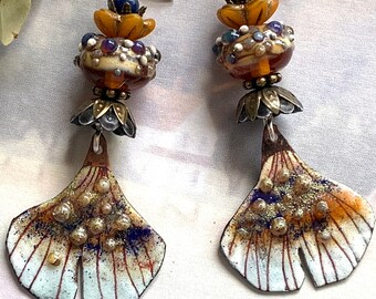 Op de afbeelding: Een paar oorhangers met een ginkgo-bladontwerp. De bladeren zijn geschilderd in tinten wit, bruin en oranje met gouden en blauwe accenten. De bovenkant van elke oorhanger heeft een cluster van amber, witte en blauwe glazen kralen.