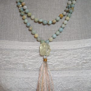 Puede incluir: Un collar de doble cadena hecho de cuentas de color azul claro y verde con un colgante de piedra tallada de color verde claro y amarillo y una borla multicolor.