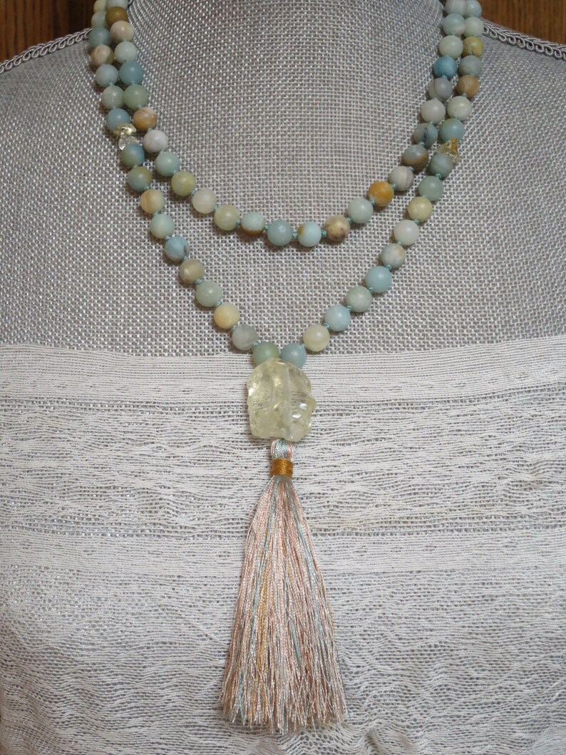 Puede incluir: Un collar de doble cadena hecho de cuentas de color azul claro y verde con un colgante de piedra tallada de color verde claro y amarillo y una borla multicolor.