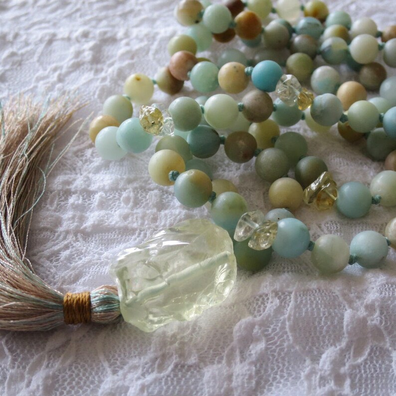Puede incluir: Un collar mala de piedras preciosas verdes y azules con un gran colgante de cristal de cuarzo transparente y un borla. El collar est&aacute; hecho de piedras naturales y est&aacute; enhebrado en un cord&oacute;n delgado.