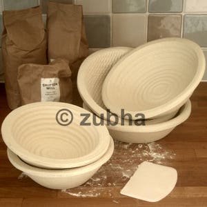 May include: Four round, woven, beige bread proving baskets of varying sizes. The baskets are on a wooden surface with flour scattered around them. A white plastic dough scraper is in the foreground.