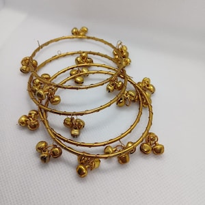 May include: Four gold-colored bangles with small, dangling bell charms. The bangles are thin and circular, with the bells evenly spaced around the circumference. The bells have a rounded shape and a shiny finish.