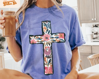 May include: A purple t-shirt with a floral cross design. The cross is outlined in black and filled with pink, orange, and green flowers.