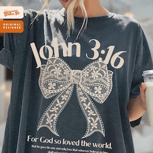 May include: Dark gray t-shirt featuring the text "John 3:16" above a large, detailed bow design. Below the bow, the shirt displays the text "For God so loved the world..." in a script font. The shirt has a relaxed fit.