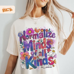 Puede incluir: Una camiseta blanca con un diseño gráfico colorido que dice "Normalizar las mentes de todo tipo". El gráfico presenta flores, libros y cerebros.