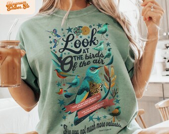 Puede incluir: Una camiseta verde con un diseño gráfico de pájaros y estrellas. El texto en la camiseta dice "Mira las aves del cielo" y "Ellos no siembran ni cosechan, ni almacenan en graneros, y sin embargo vuestro Padre celestial los alimenta. ¿No valéis vosotros mucho más que ellos? Mateo 6:26-27".