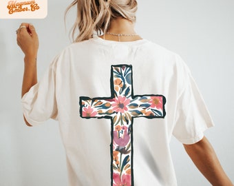May include: White t-shirt with a floral cross design on the back. The cross is outlined in black and filled with pink, orange, and green flowers.
