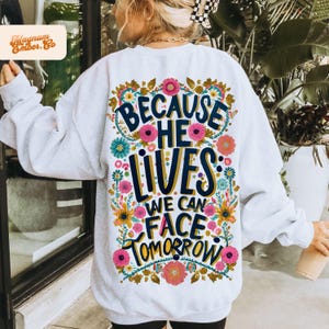 Puede incluir: Sudadera blanca con un diseño floral y el texto "Because He Lives: We Can Face Tomorrow".