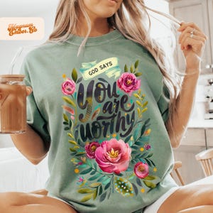 Puede incluir: Una camiseta verde con un diseño floral y el texto "God Says You Are Worthy".
