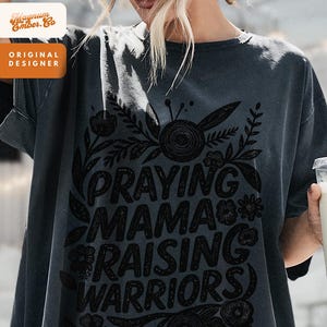 May include: Dark gray t-shirt with the text "PRAYING MAMA RAISING WARRIORS" in a black floral design. The shirt also has the text "Magnum Ember. Co" and "ORIGINAL DESIGNER" in a white and orange design.