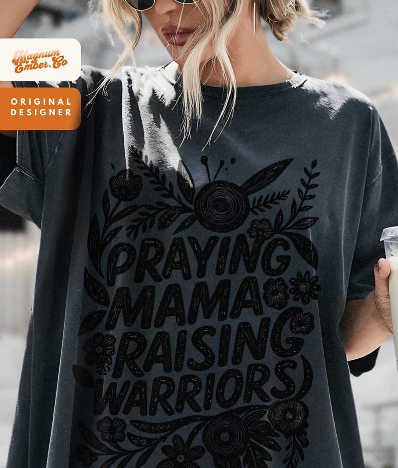 May include: Dark gray t-shirt with the text "PRAYING MAMA RAISING WARRIORS" in a black floral design. The shirt also has the text "Magnum Ember. Co" and "ORIGINAL DESIGNER" in a white and orange design.