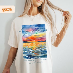 May include: White t-shirt with a watercolor painting of a sunset over a body of water. The text "deep waters" is written in blue cursive above the painting. The text "WHEN YOU GO THROUGH DEEP WATERS I WILL BE WITH YOU... Isaiah 43:2-3" is written in blue below the painting.